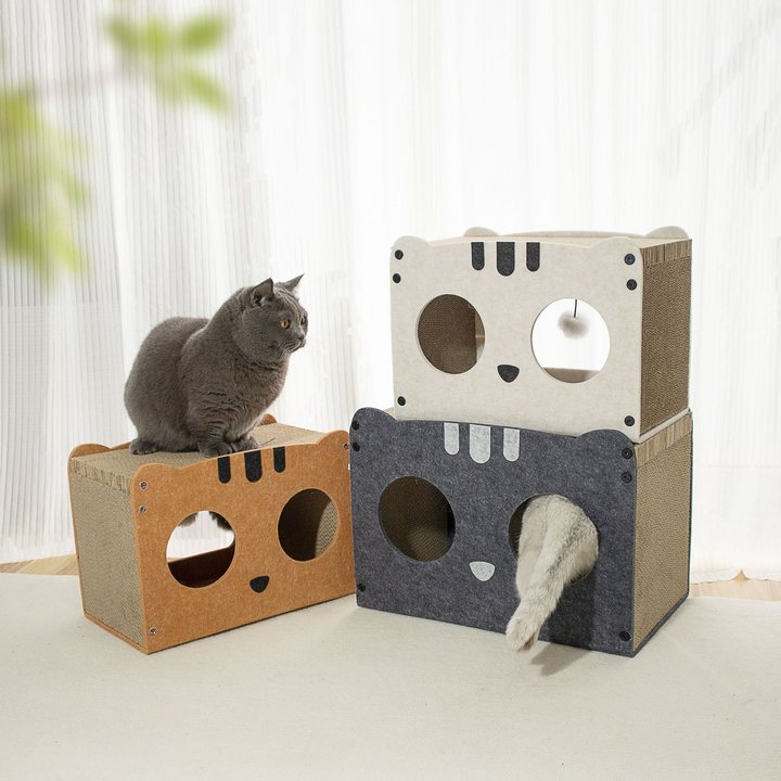 Stacked cat-face shaped cardboard cat houses with round entry holes, one grey cat sitting on top near sheer curtains