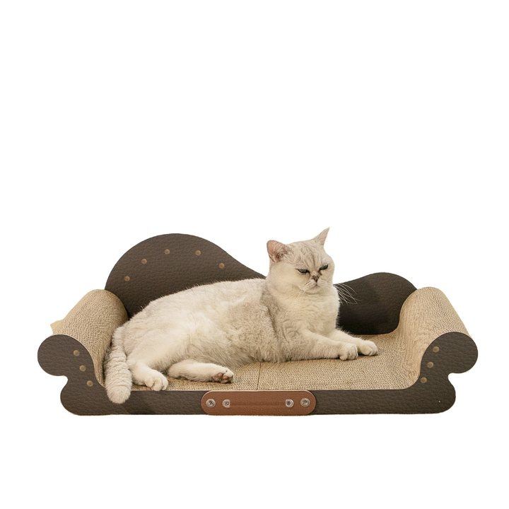 A scalloped, Victorian-style pet lounge/scratcher shaped like a miniature sofa, made of corrugated cardboard with a beige woven-texture seat/base and dark brown padded backrest and armrests accented with small round studs; a cream-and-grey British Shorthair cat lies stretched across it.